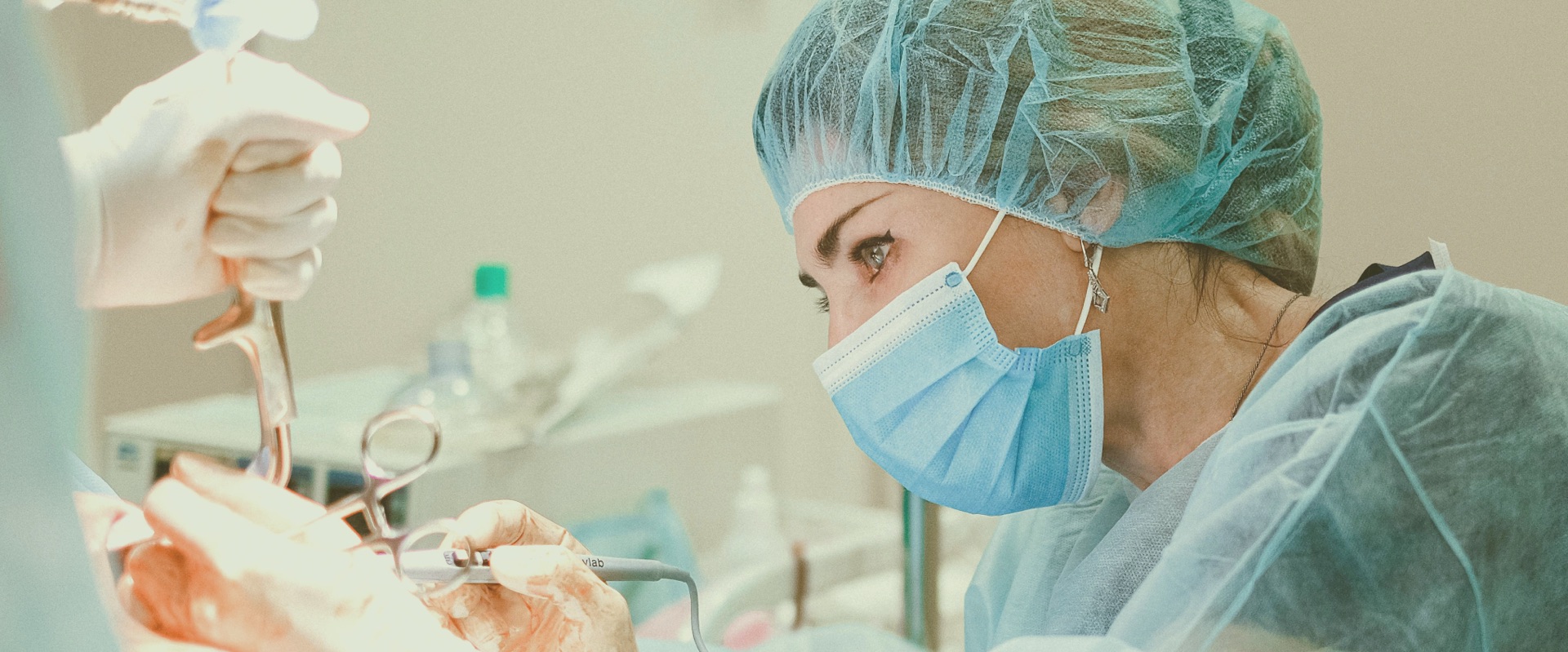Surgeon in blue surgical mask and hair cap performing a procedure with surgical instruments, with medical equipment and bright surgical lighting visible in the background